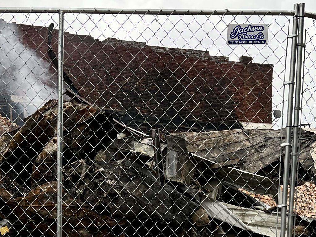 Burned debris behind a chain-link fence, with smoke rising from a damaged structure
