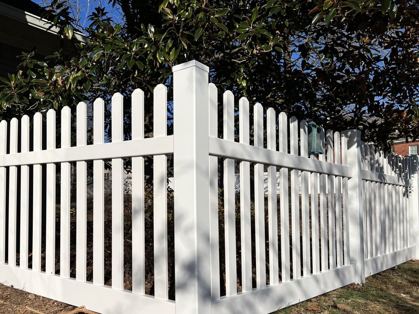 White wooden picket fence with a corner post in front of trees and a house in sunlight