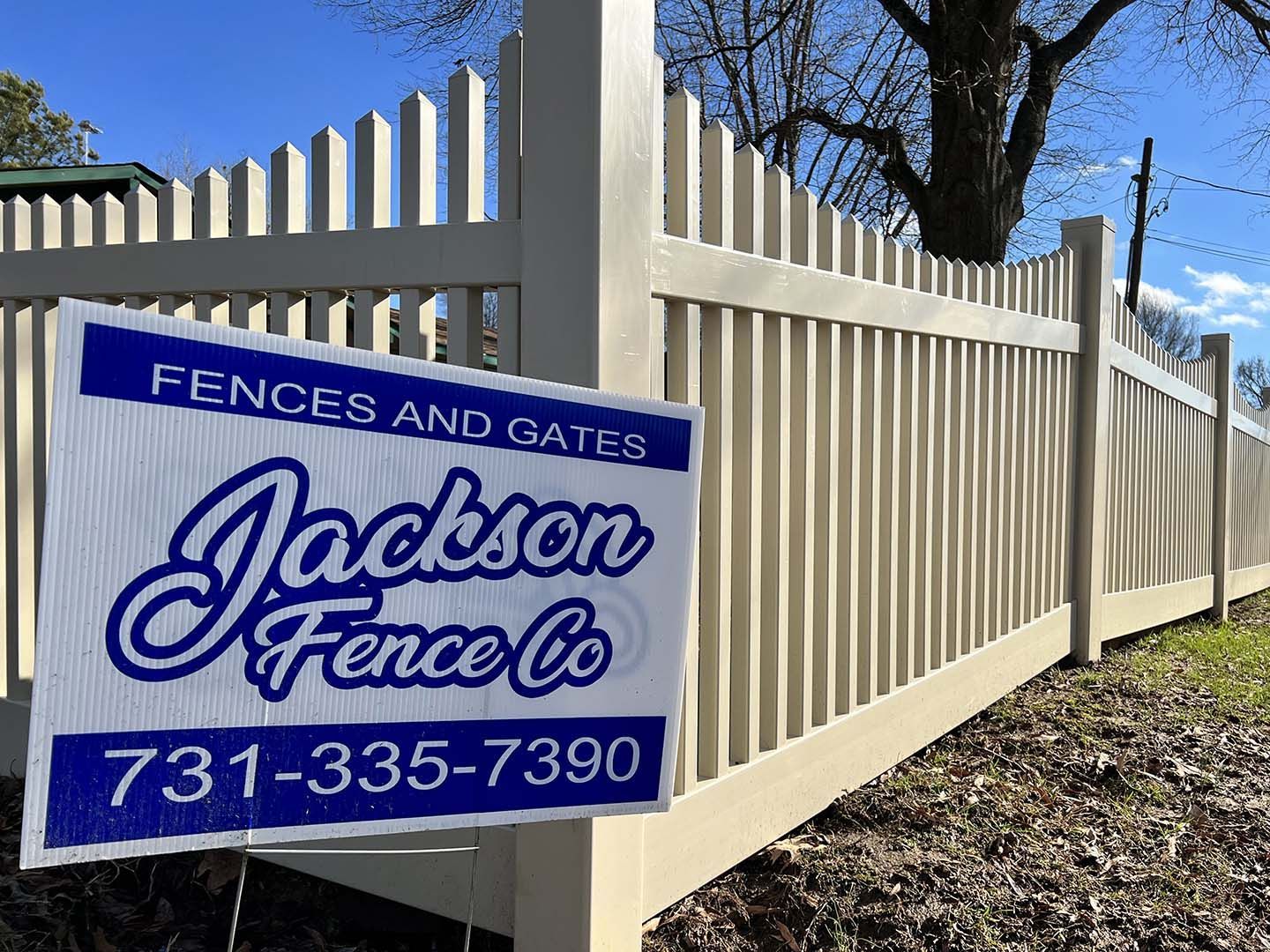 Blue fence and gate sign for Jackson Fence Co. along a tan vinyl fence under a clear sky.