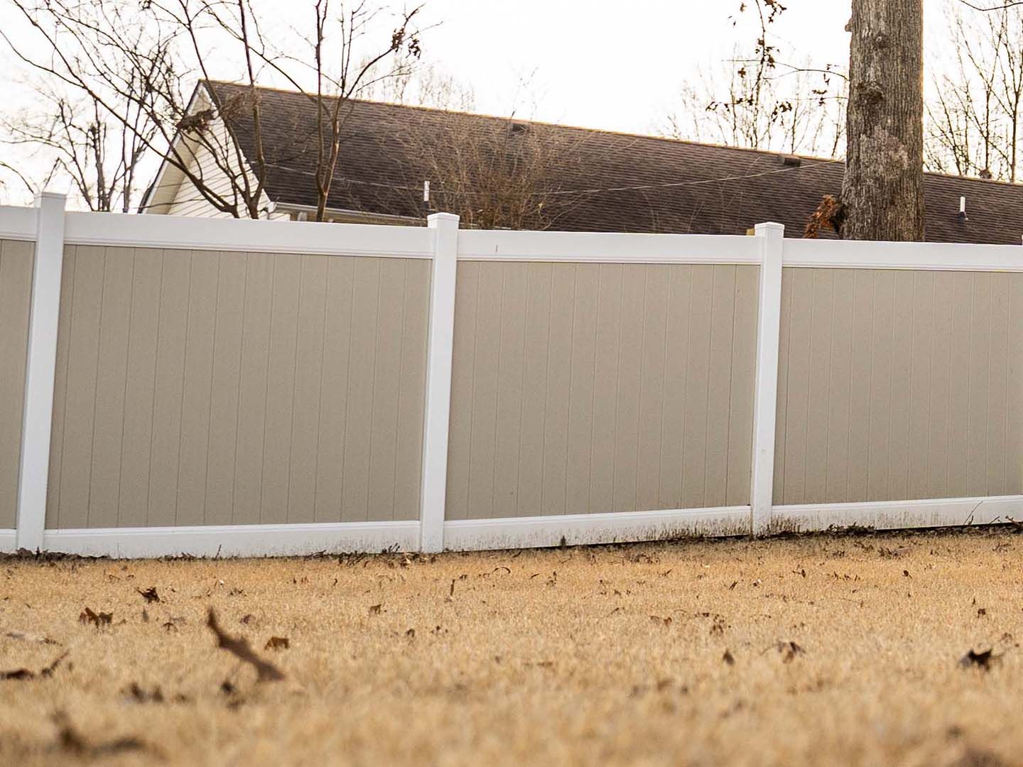 Beige privacy fence with white posts in a yard, with a house and trees behind it