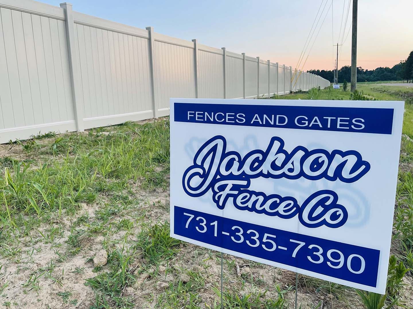 Jackson Fence Co. roadside sign by a white fence at dusk, blue sign with phone number 731-335-7390