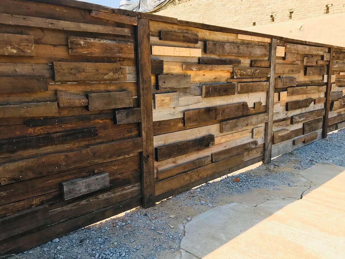 Wooden slat fence in mixed brown tones along a gravel path beside a light-colored wall