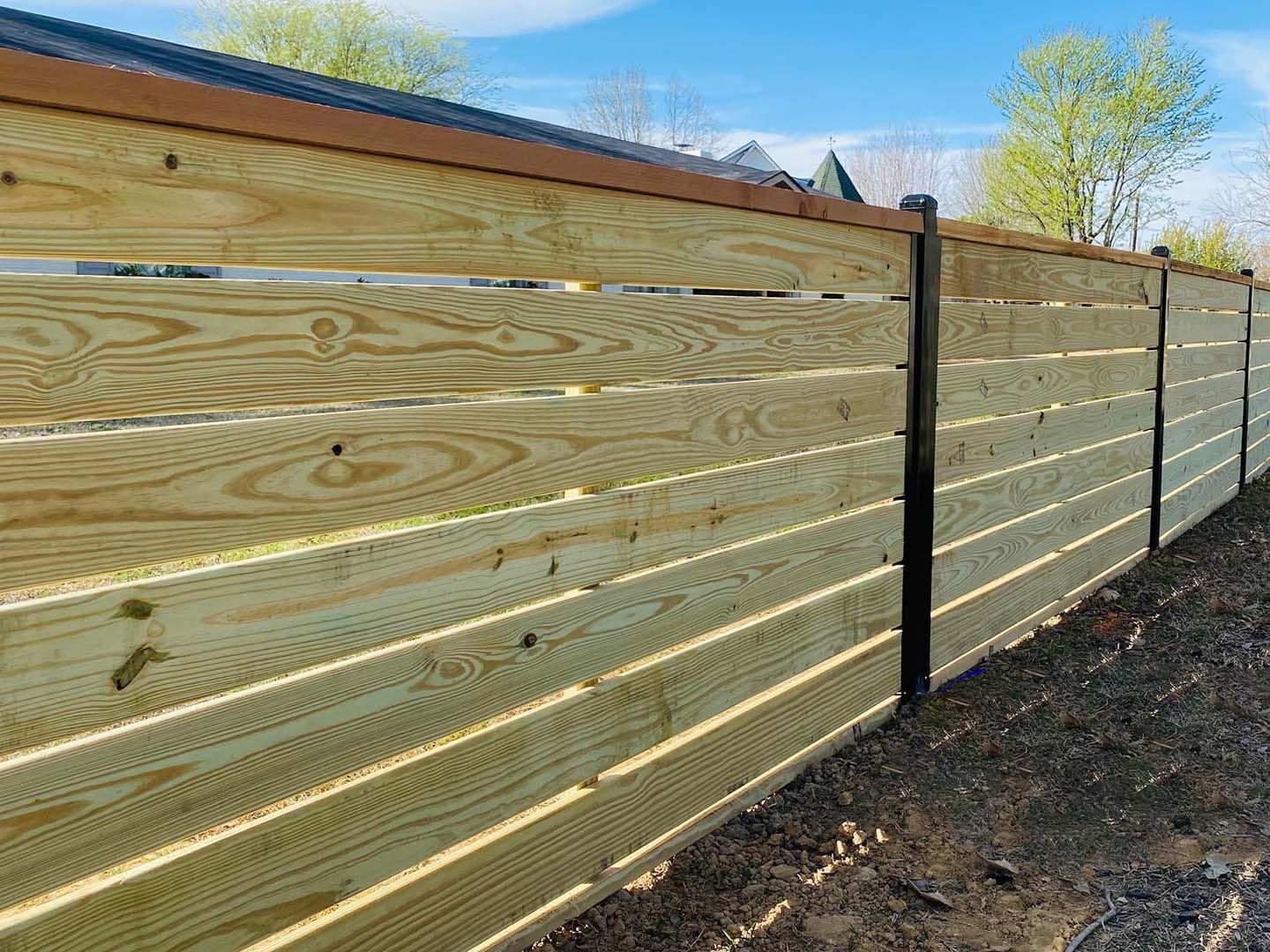 Wooden privacy fence with horizontal slats and black posts along a dirt yard under a blue sky