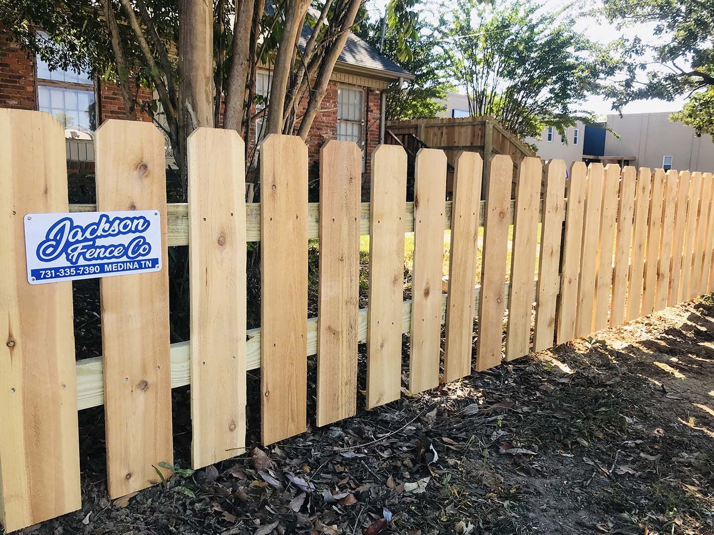 Wooden picket fence with a blue sign in a yard, beside trees and a house
