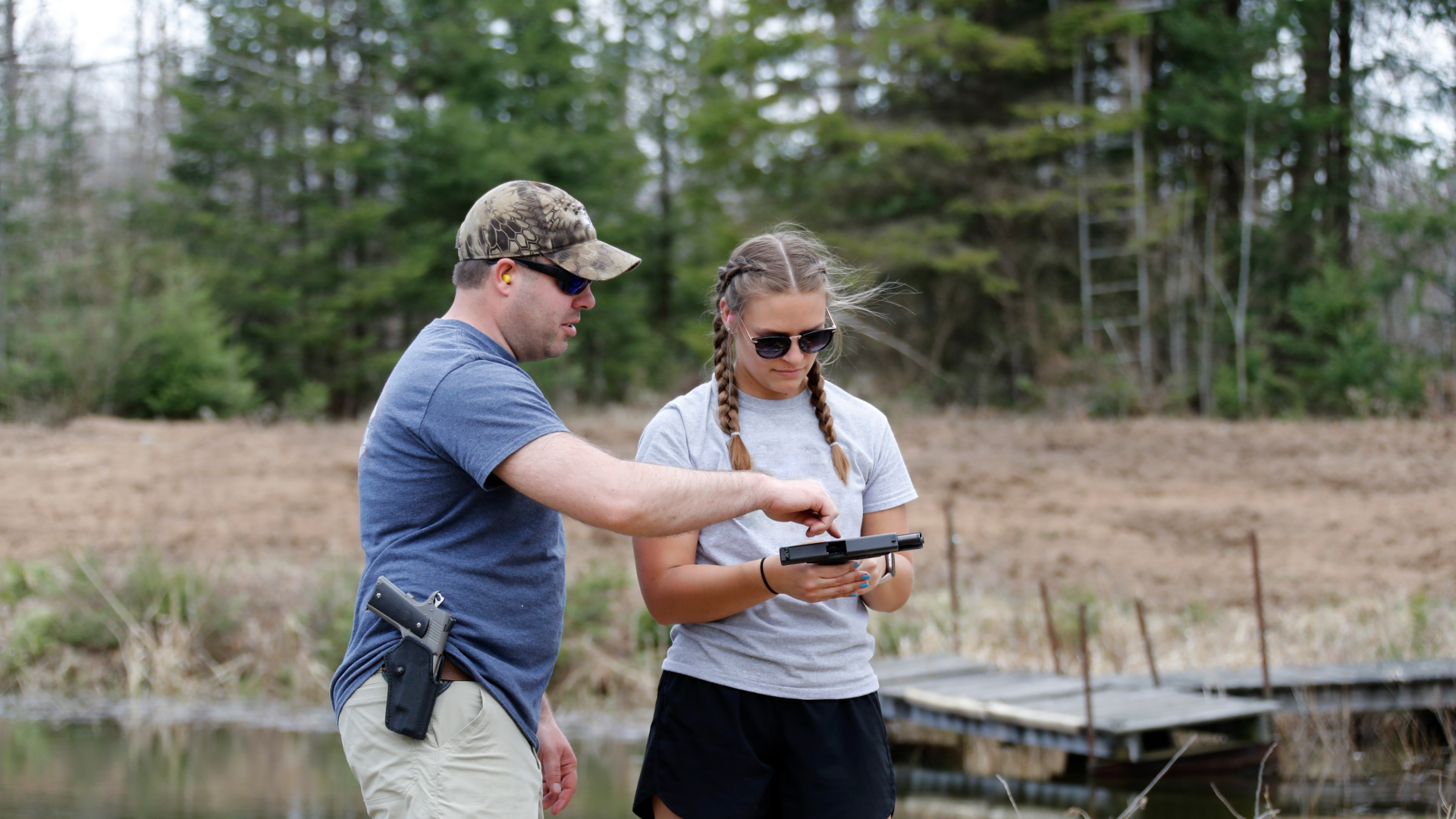 Woman at a shooting range smiles near a target with bullet holes. Blue sky, outdoor setting.