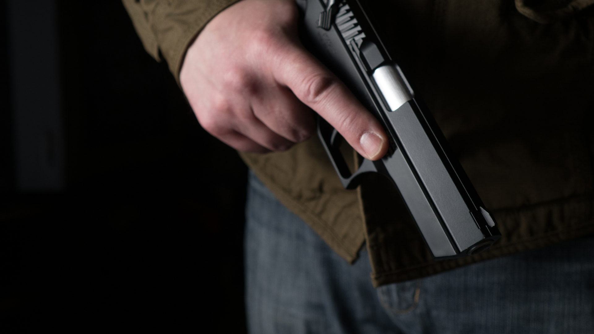 Close-up of a hand holding a handgun beside camouflage pants in a bright indoor hallway.