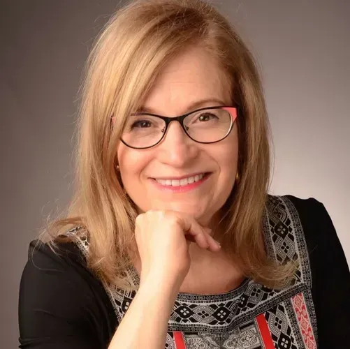 A woman wearing glasses and a black shirt is smiling with her hand on her chin.