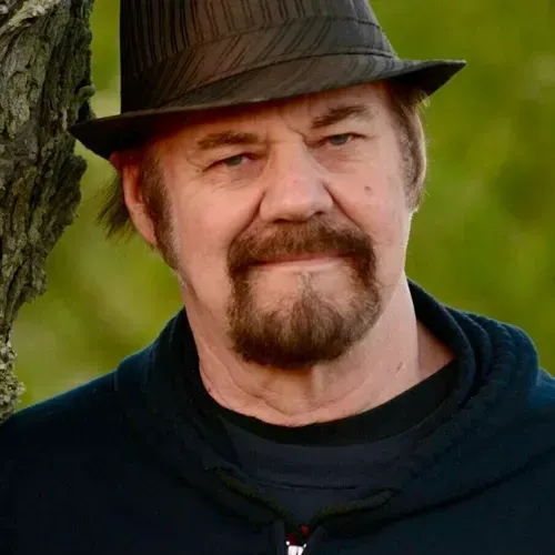 A man with a beard and hat is leaning against a tree.