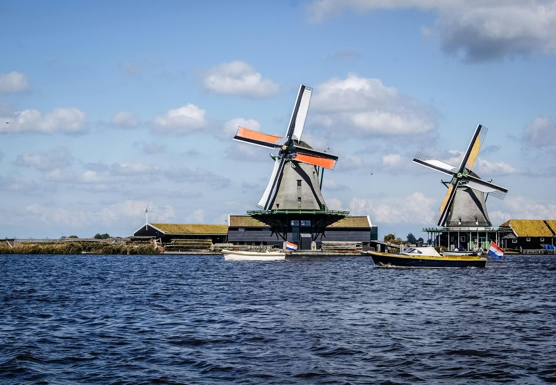 Two windmills with orange and white sails, overlooking a body of water, boats present; Netherlands.