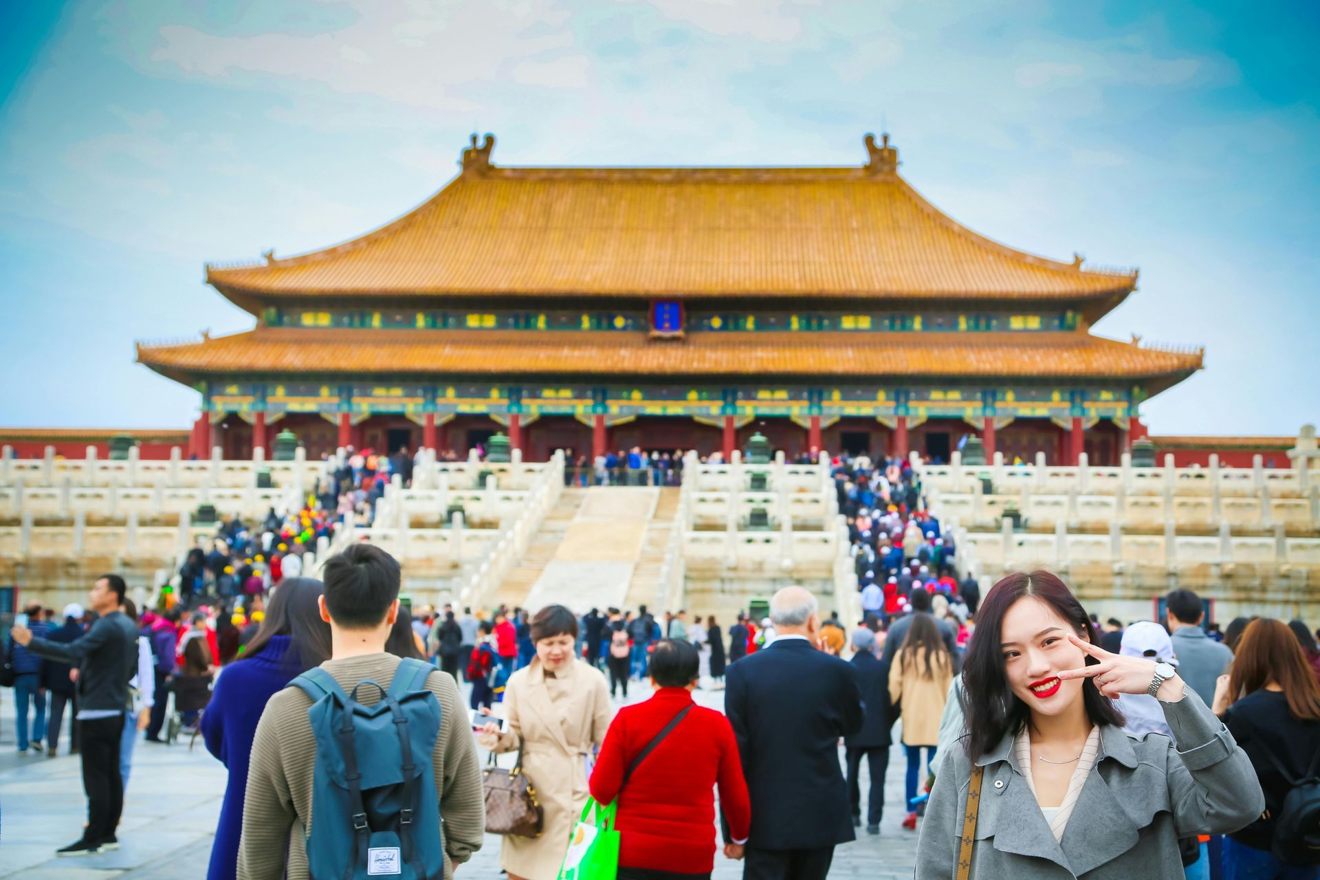 Woman making a hand gesture smiles in front of the Forbidden City in Beijing, China; tourists fill the courtyard.