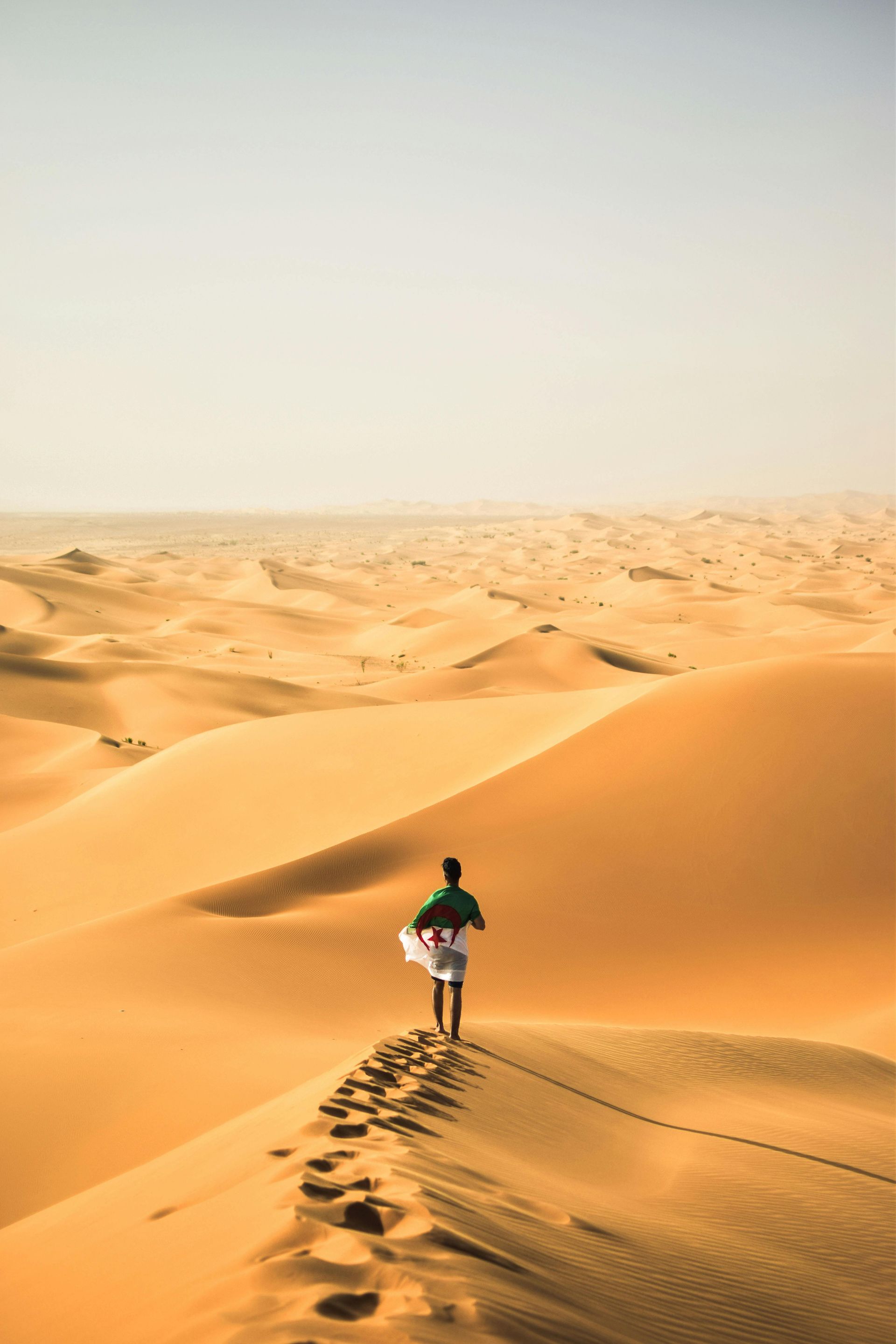 Person walking on a sand dune in a desert, carrying a white cloth, leaving footprints.