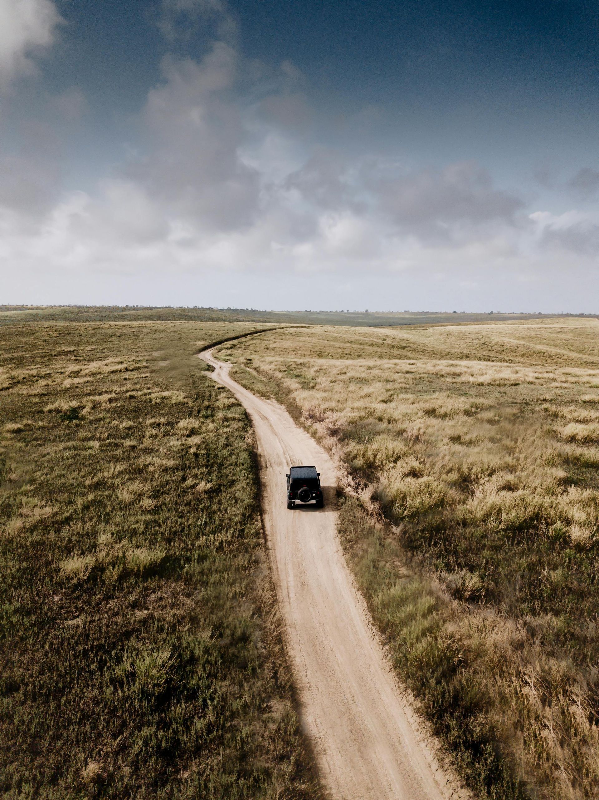 SUV driving on a dirt road through a grassy field, under a cloudy blue sky.