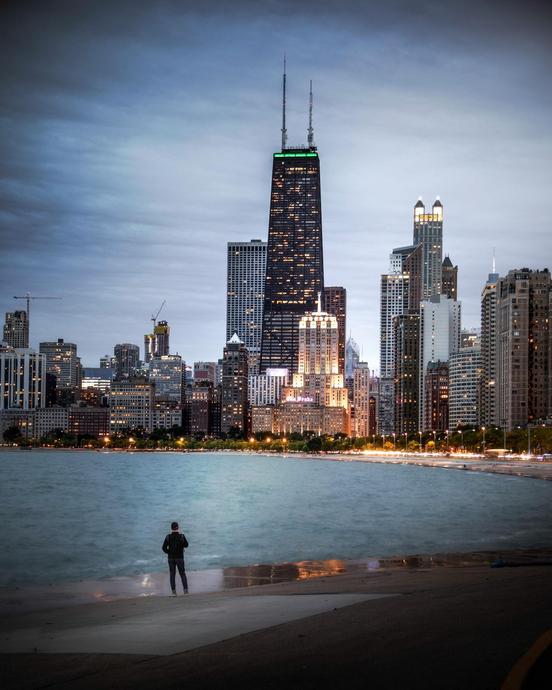 Person standing on pier, Chicago skyline at dusk. Buildings lit up against cloudy sky; water in foreground.