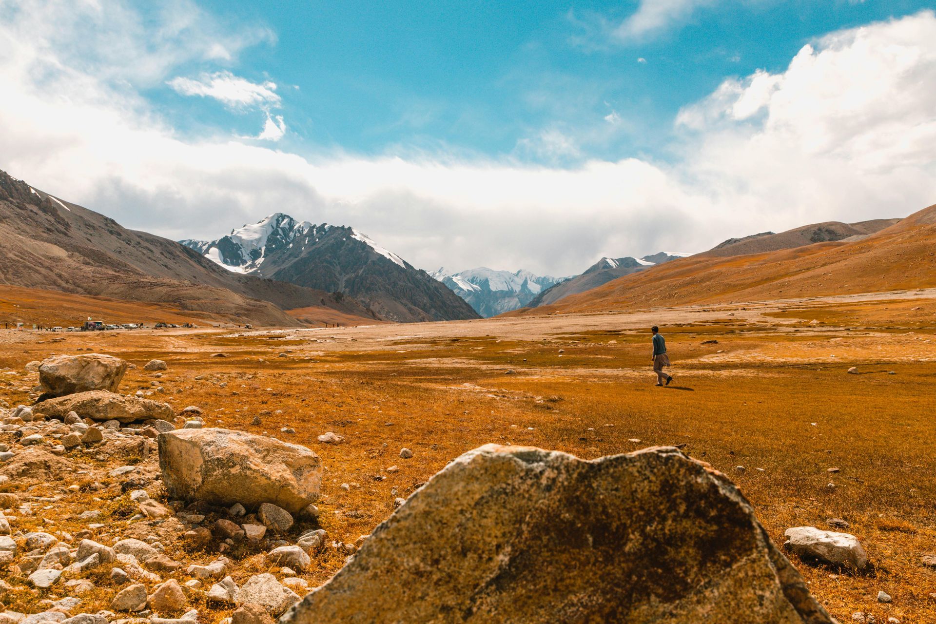 A person walks in a wide valley with mountains in the distance under a blue sky with clouds.
