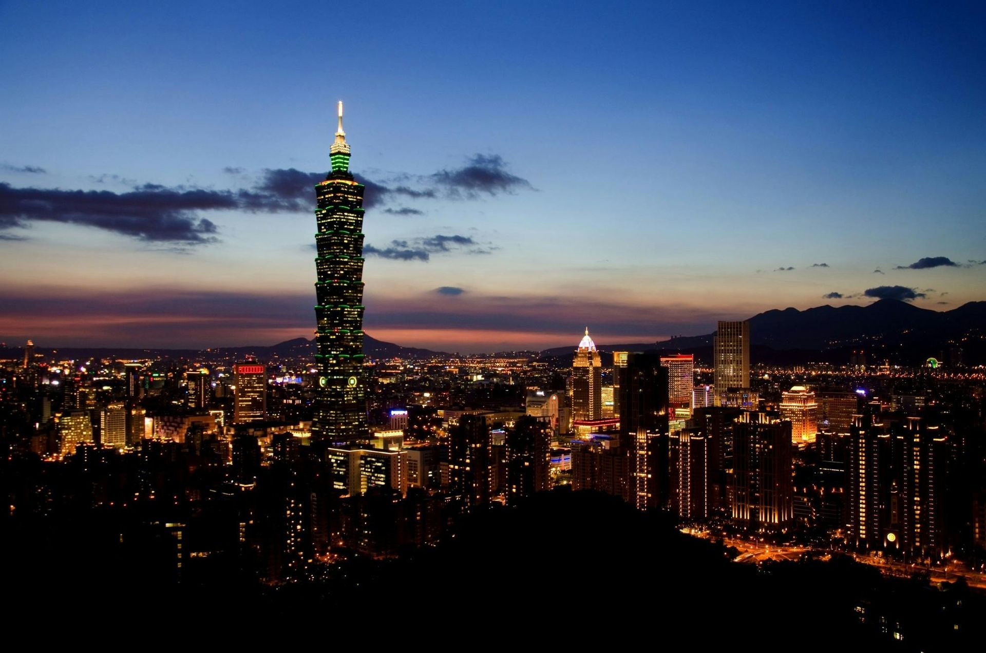 City skyline at dusk, lit buildings, Taipei 101 tower prominent against dark blue sky.