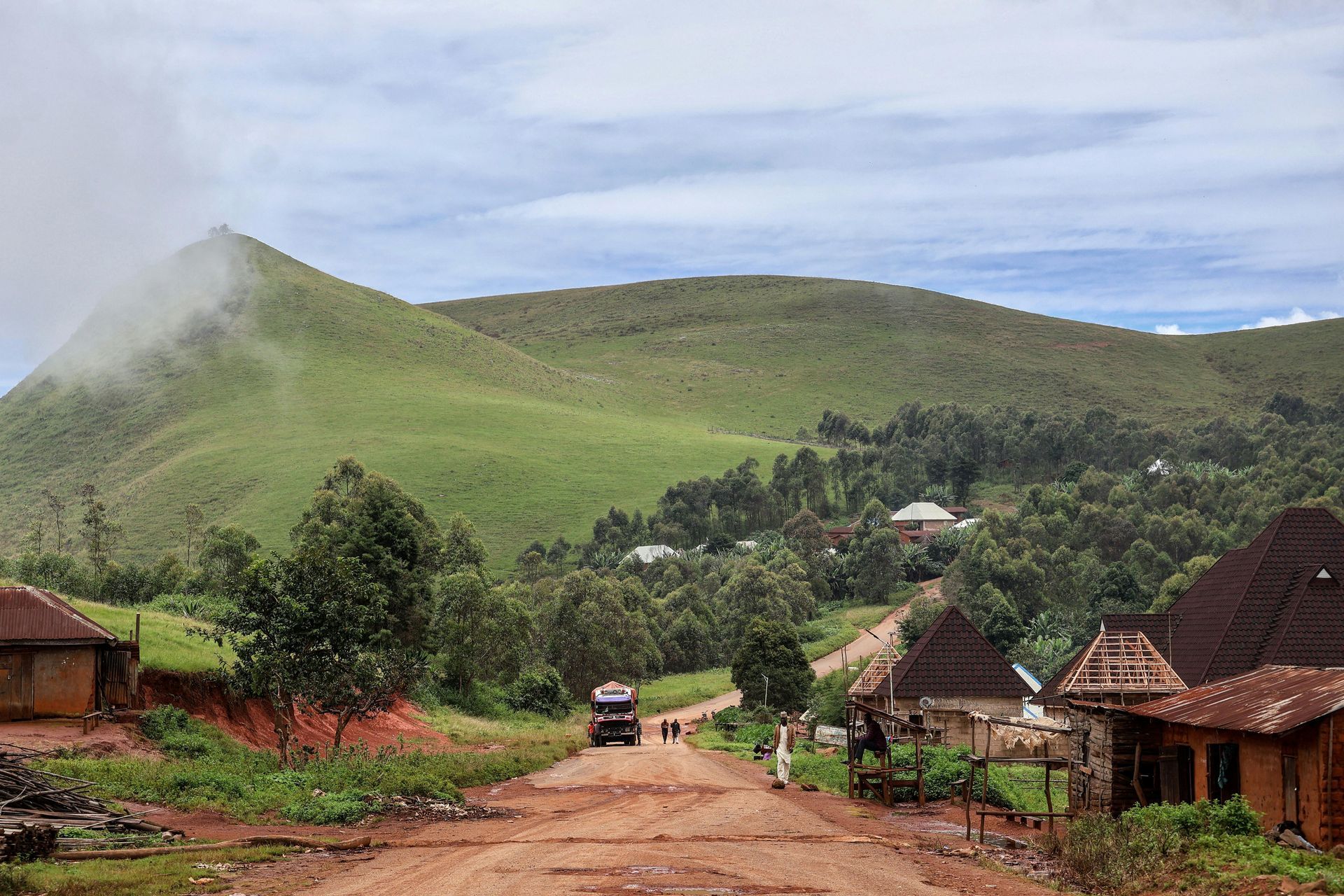 Dirt road through village leading to green hills under cloudy sky.