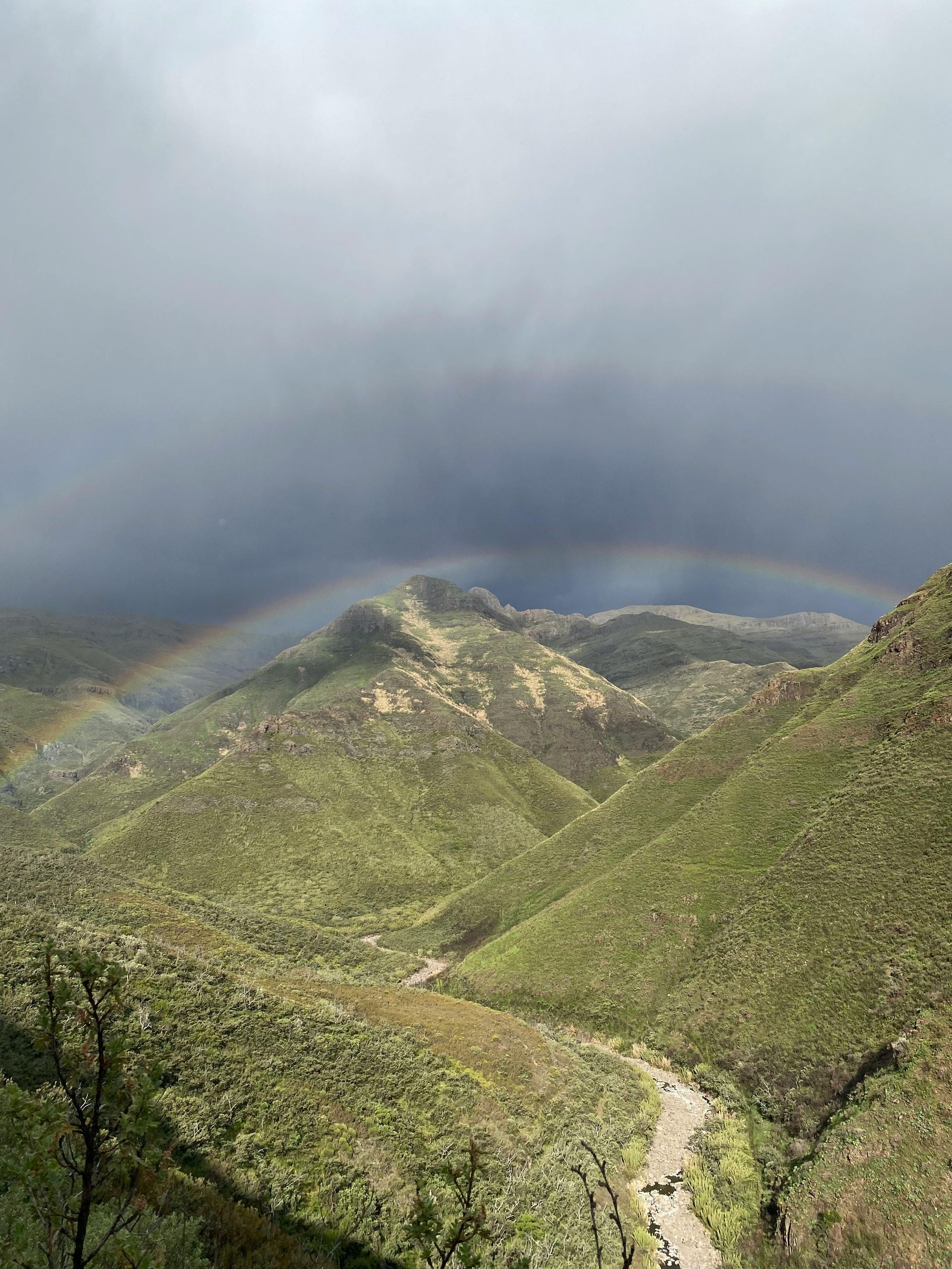 Rainbow arcs over green mountains with a small river below, under a dark, stormy sky.