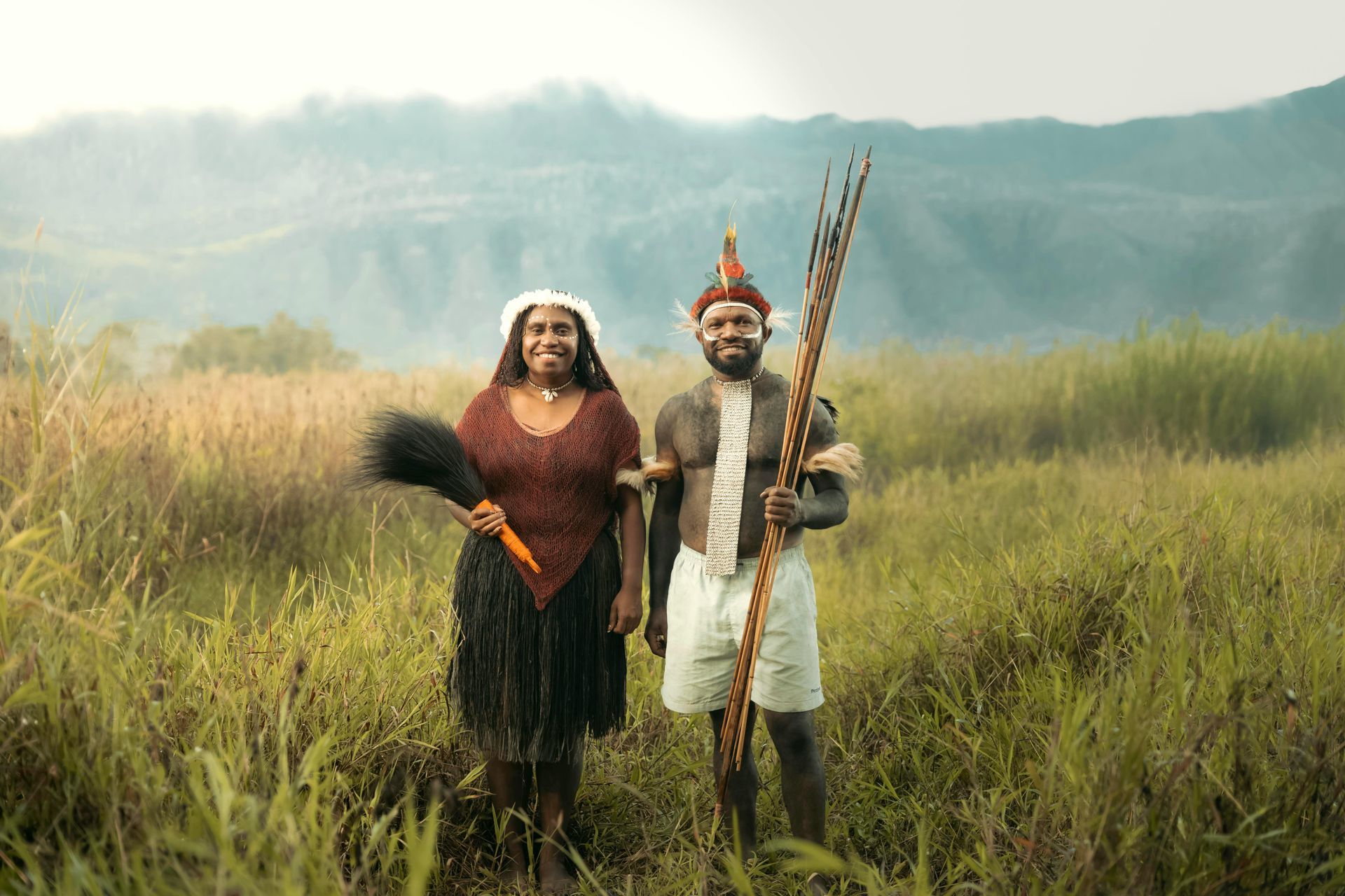 Two people in traditional attire stand in a field. Mountain backdrop. Woman holds a plume, man holds a bundle.