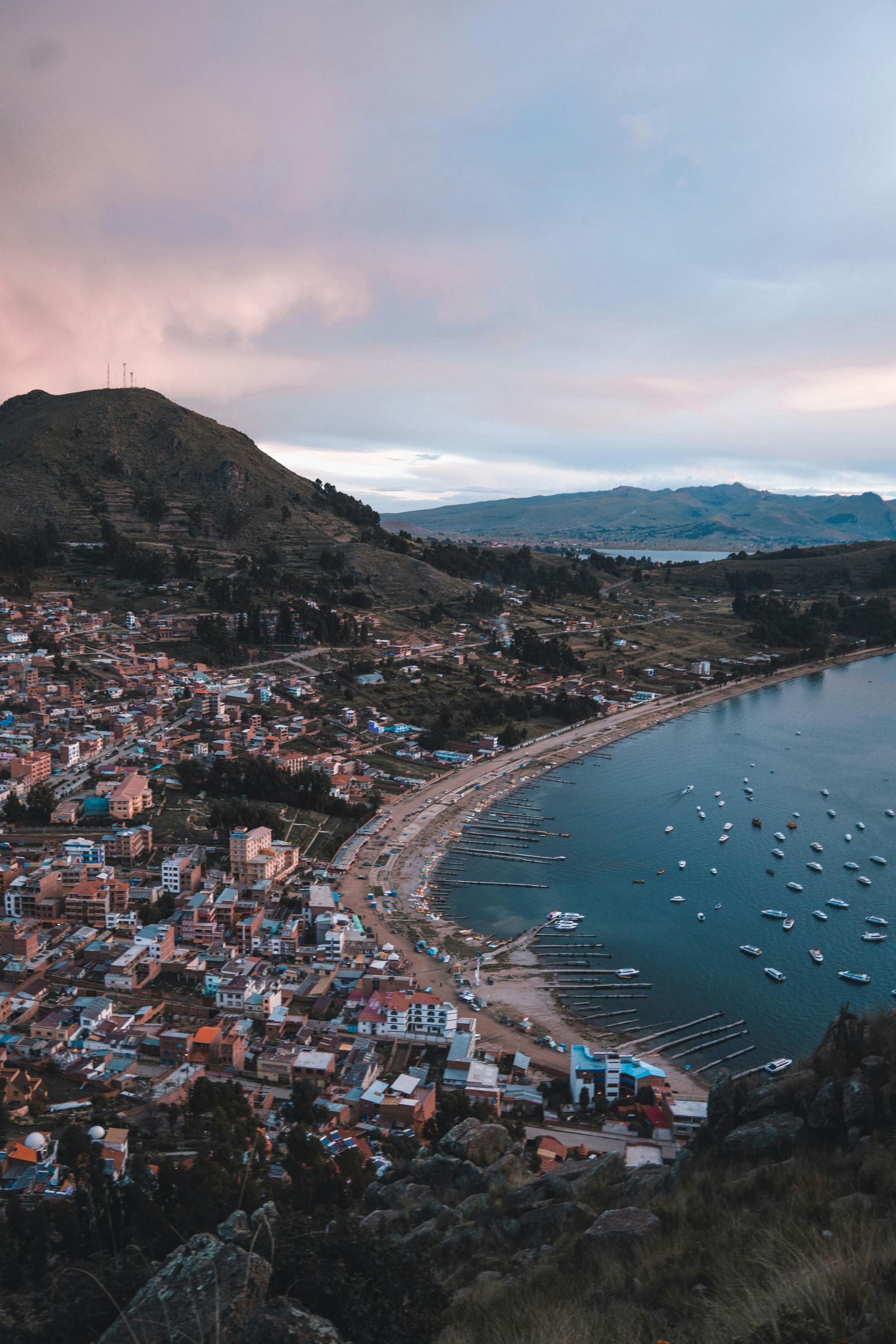 Coastal city nestled by a bay under a dusky sky; buildings line the shore, boats dot the water, mountain in the background.