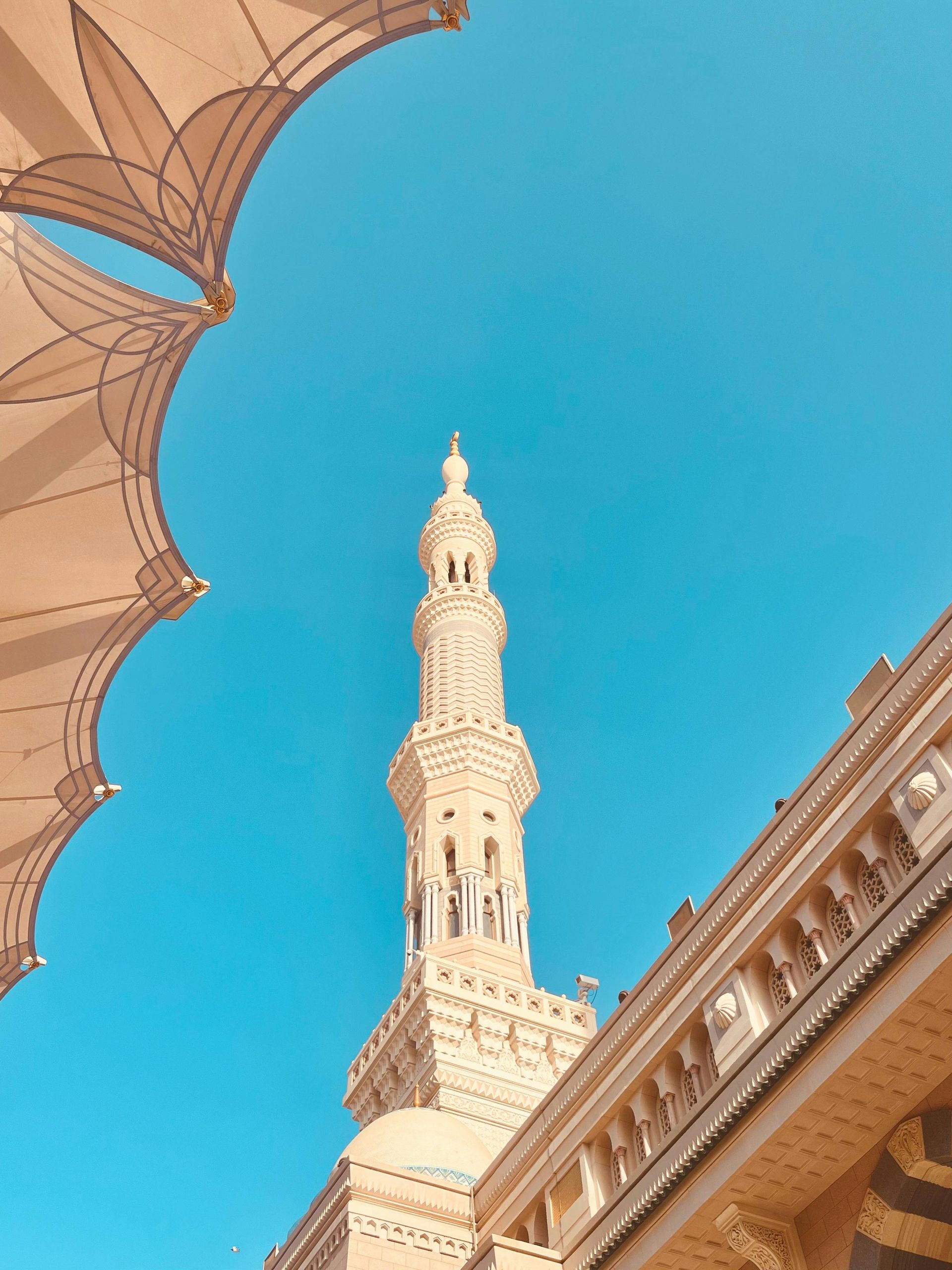 Minaret of a mosque against a bright blue sky, viewed from beneath an ornate awning.