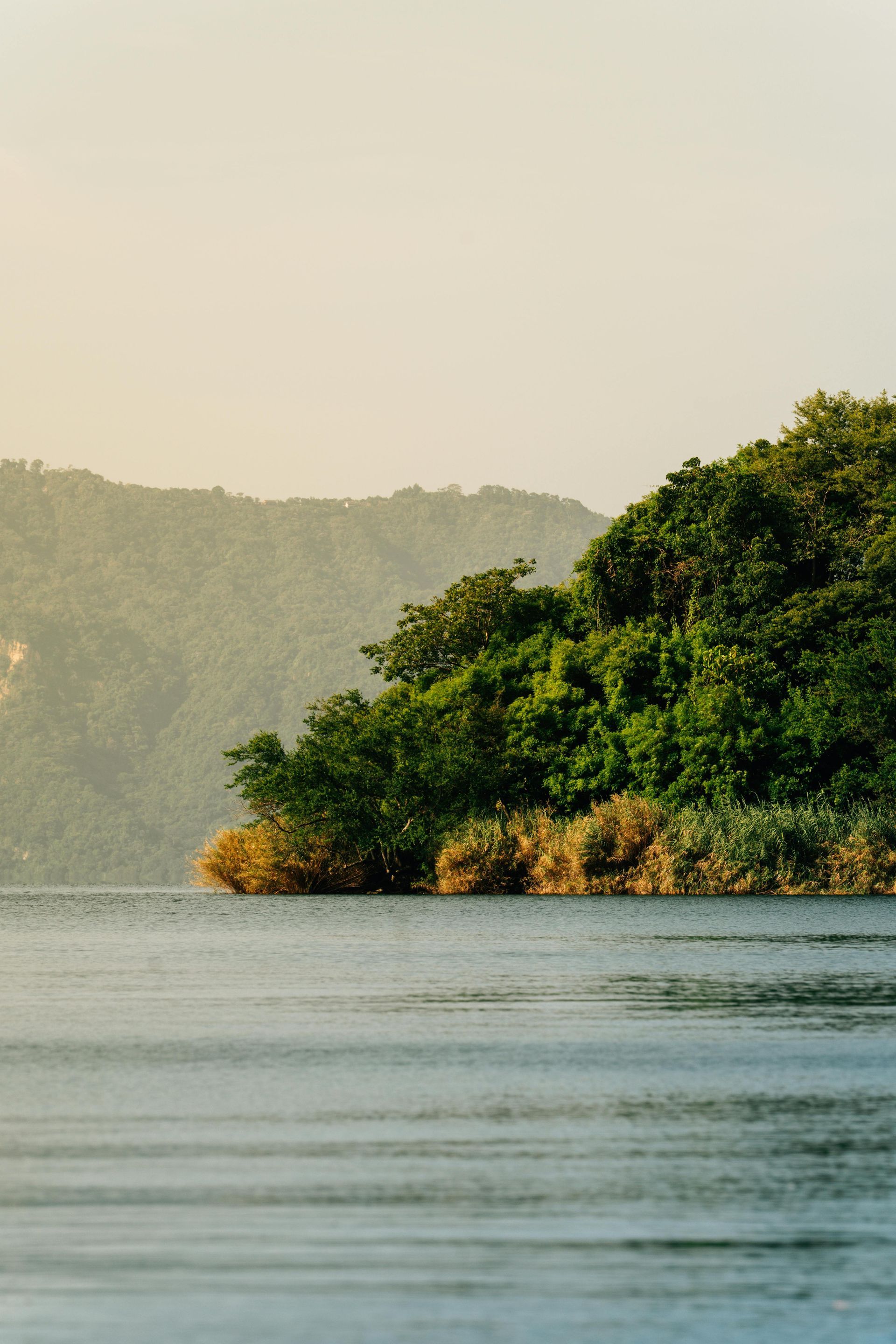 Lake with lush, green shoreline against a hazy mountain backdrop. Calm, blue water in the foreground.