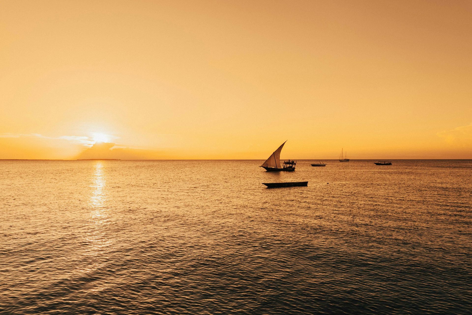 Golden sunset over calm ocean with boats and a sailing dhow silhouetted.