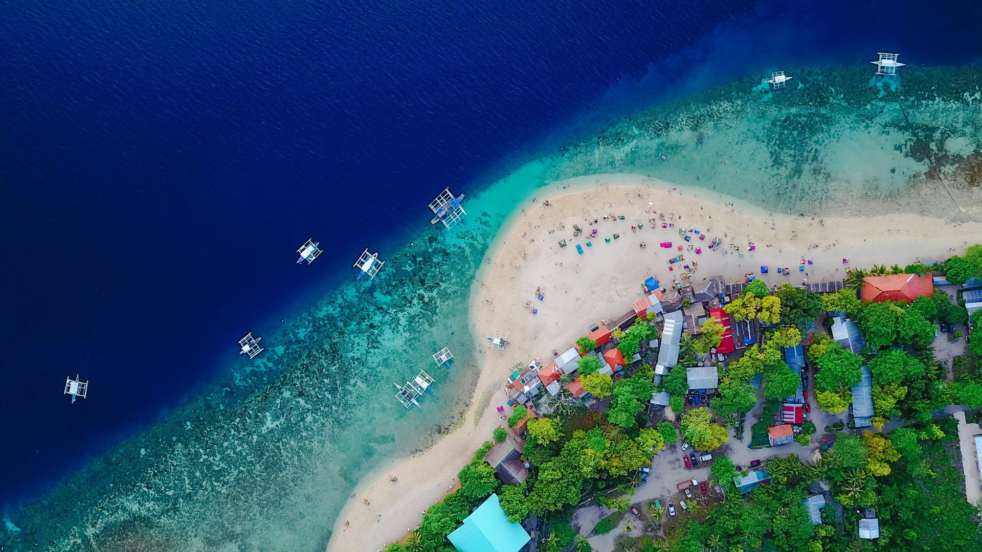 Aerial view of a beach with turquoise shallows meeting deep blue ocean. Boats and a village line the shore.