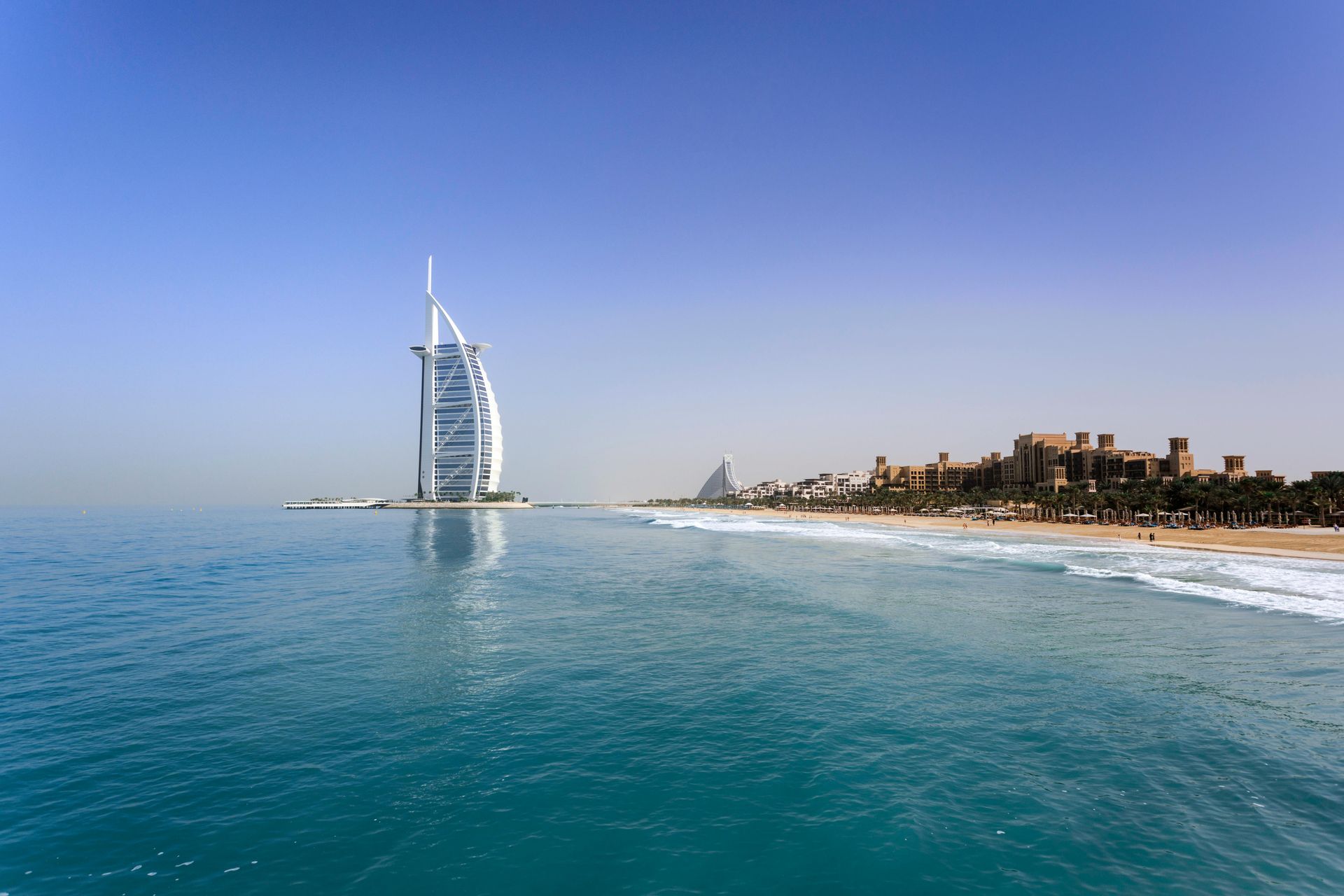 Burj Al Arab hotel in Dubai, seen from the ocean, with a beach and clear blue sky.