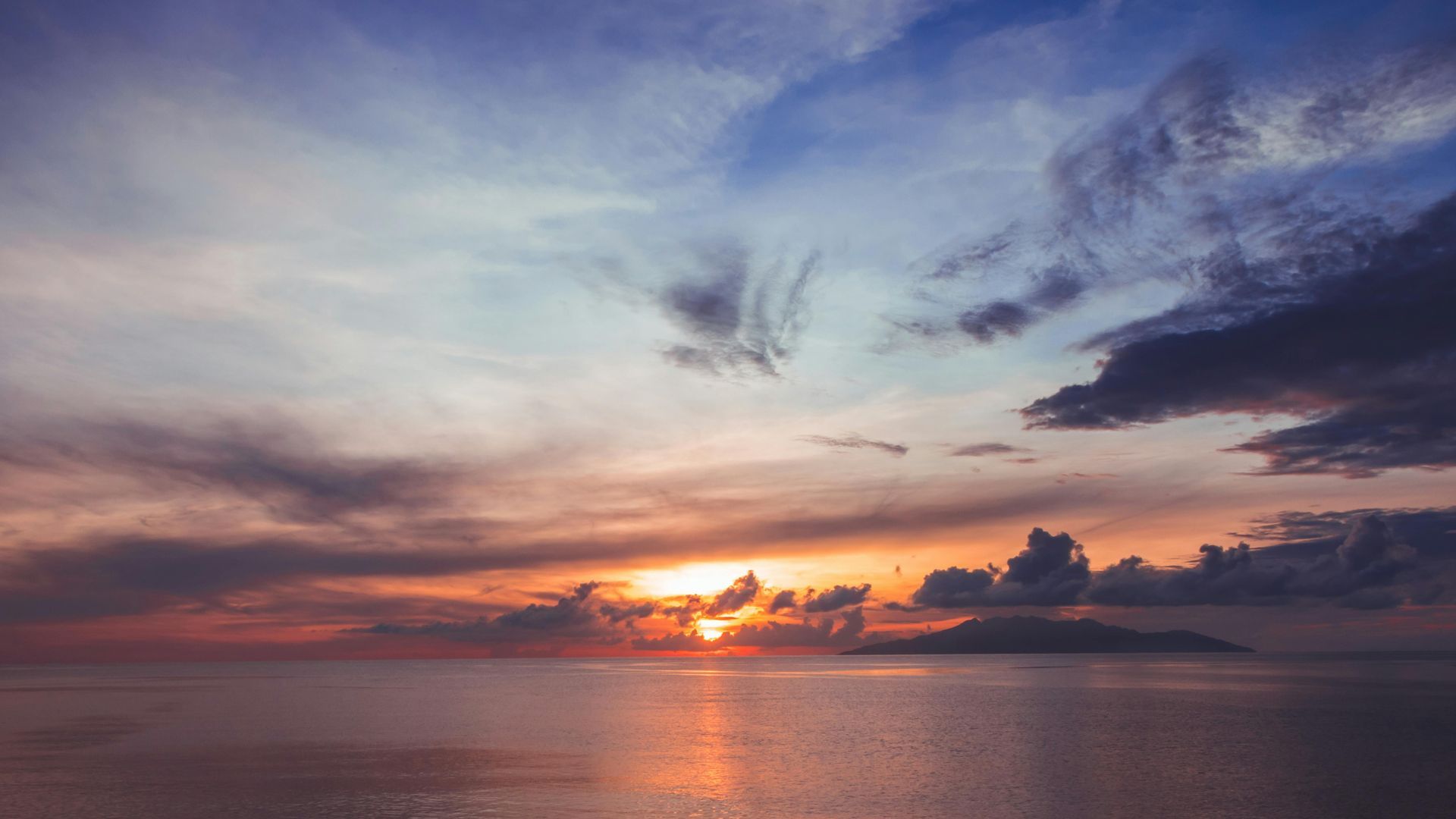 Sunset over calm ocean, orange and blue sky, dark clouds.
