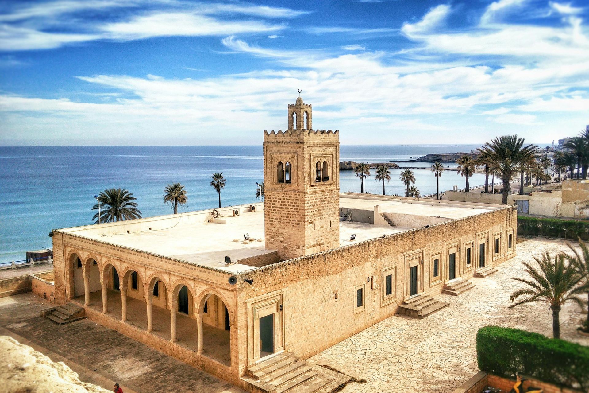 Mosque with a minaret by the sea, palm trees, and blue sky. Beige building.