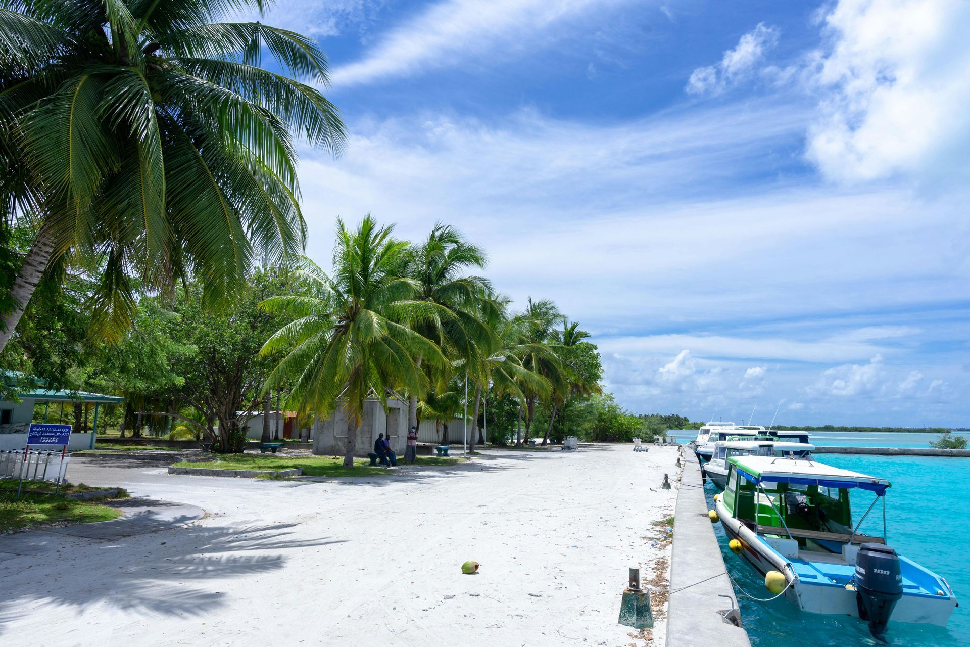 Palm trees line a white sandy beach with turquoise water. Boats docked at a pier under a blue sky.