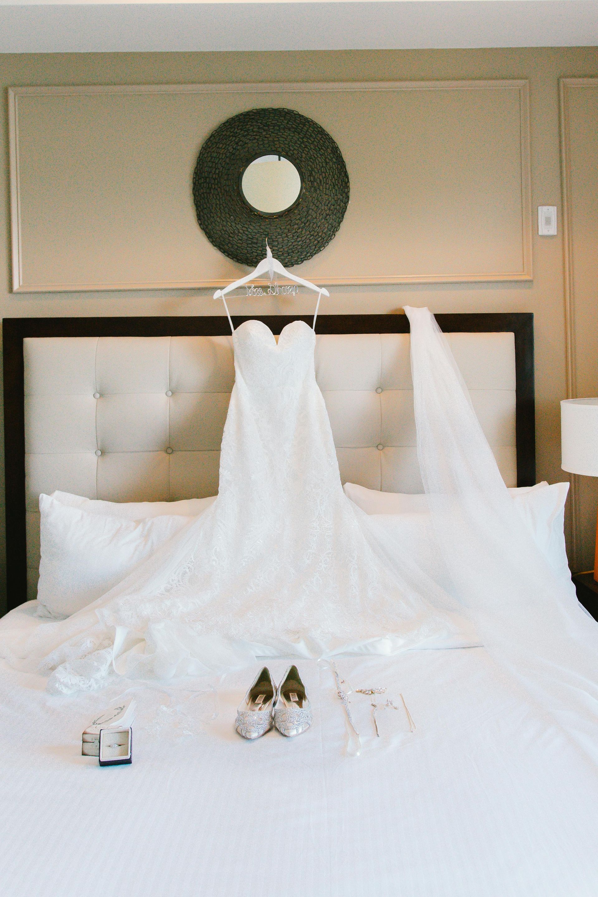 Wedding dress and veil on a bed, with shoes and accessories laid out; a decorative mirror hangs above.