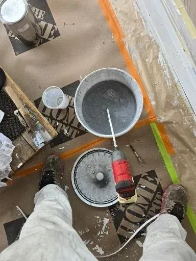 Overhead view of paint buckets and tools on a floor beside an orange-marked wall, with a person’s shoes visible