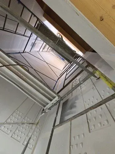 Open elevator shaft with metal beams and safety rails, viewed upward from inside a building.