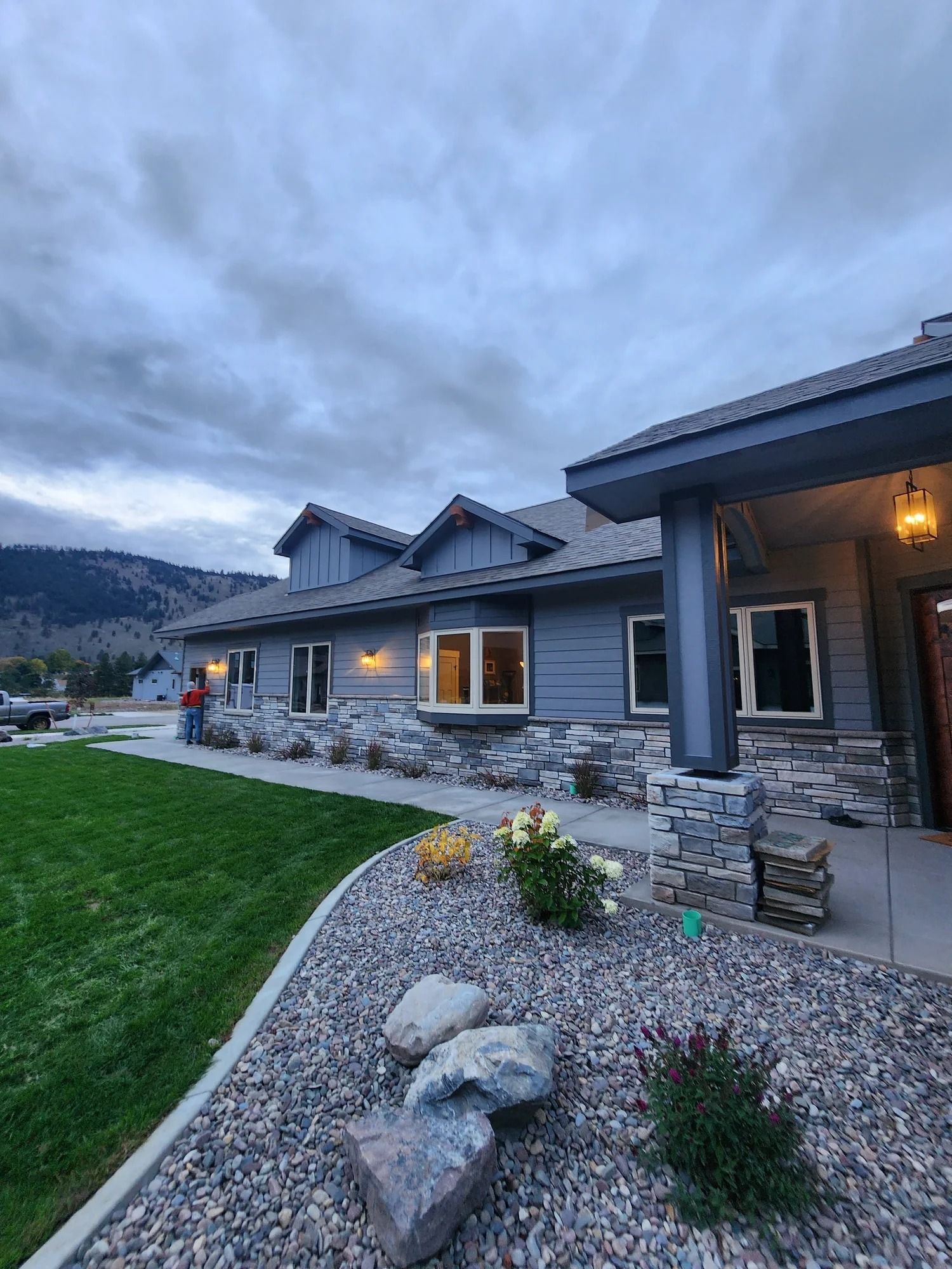 Modern stone house at dusk with lit windows, gravel landscaping, and mountains in the background