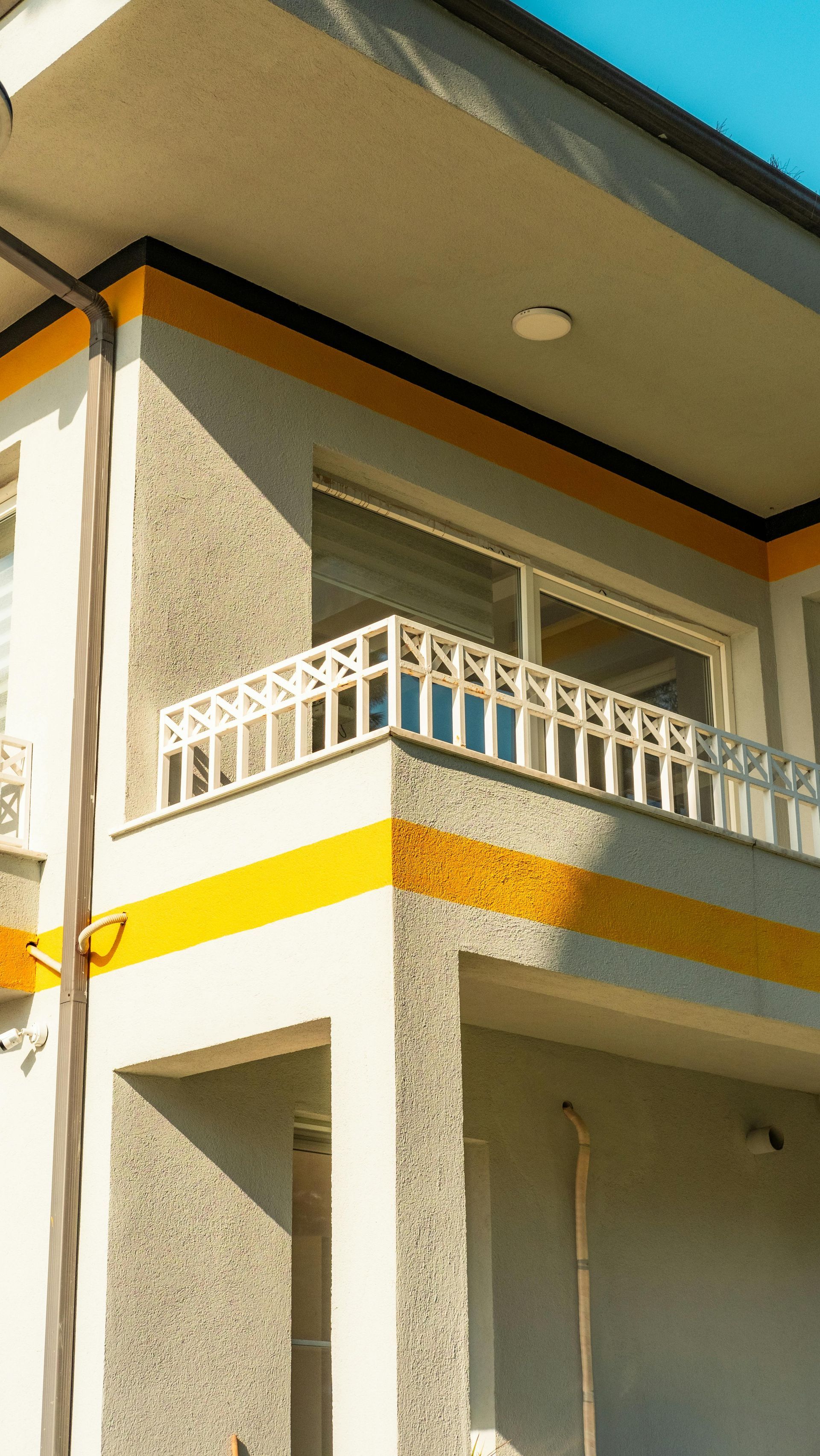 Exterior corner of a beige house with white balcony railing and orange trim under a blue sky