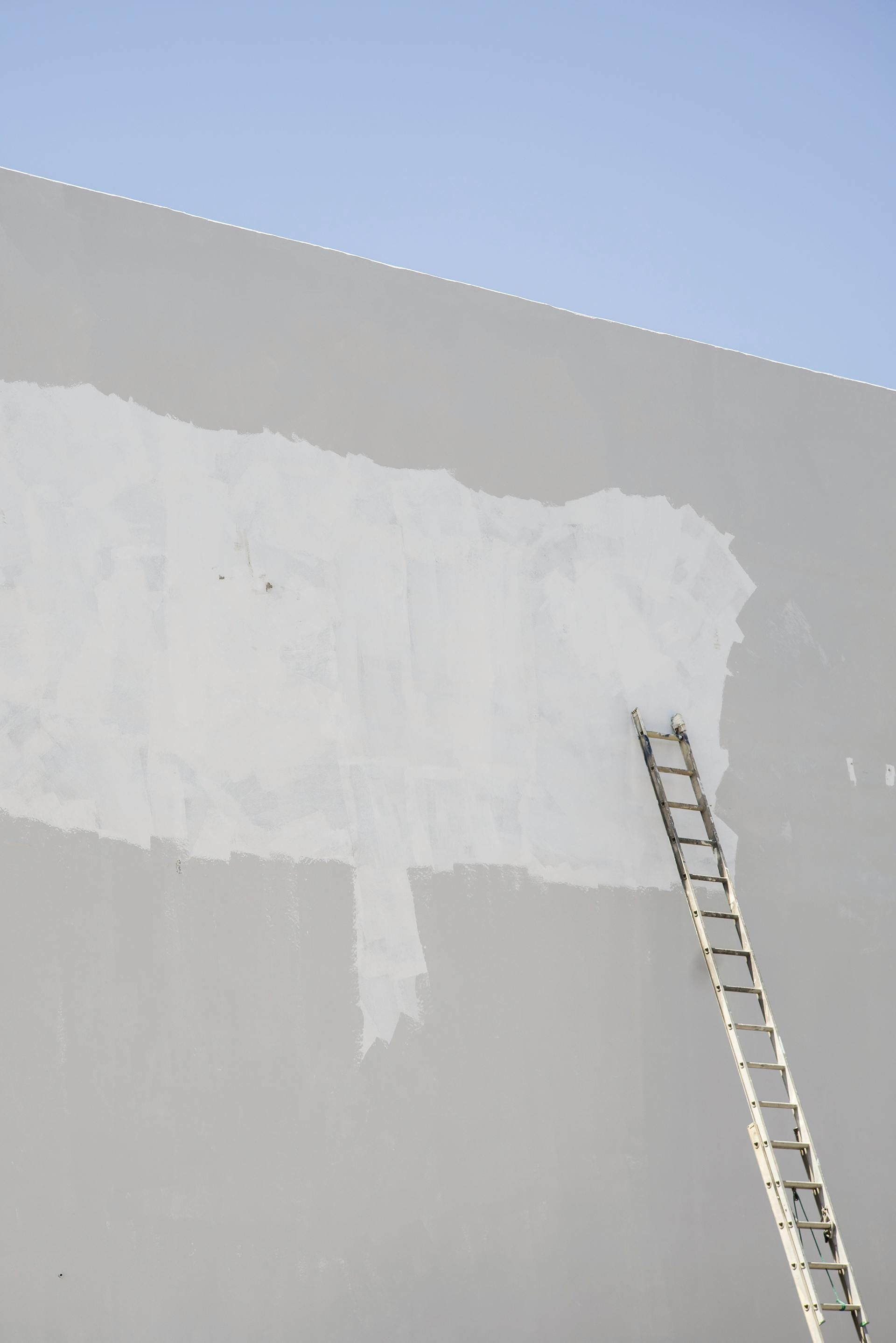 Ladder leaning against a gray wall with a large white painted patch under a blue sky