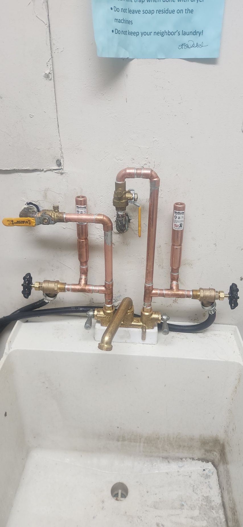 Plumbing pipes and valves mounted on a wall above a white sink, with a blue note pinned above.
