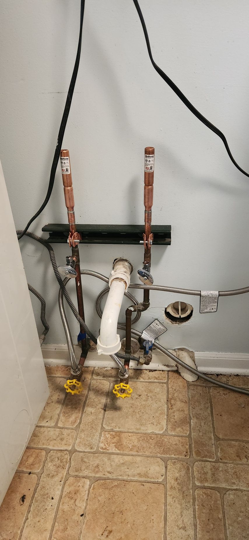 Plumbing pipes and valves against a wall, with a white sink or toilet pipe on a tiled floor.
