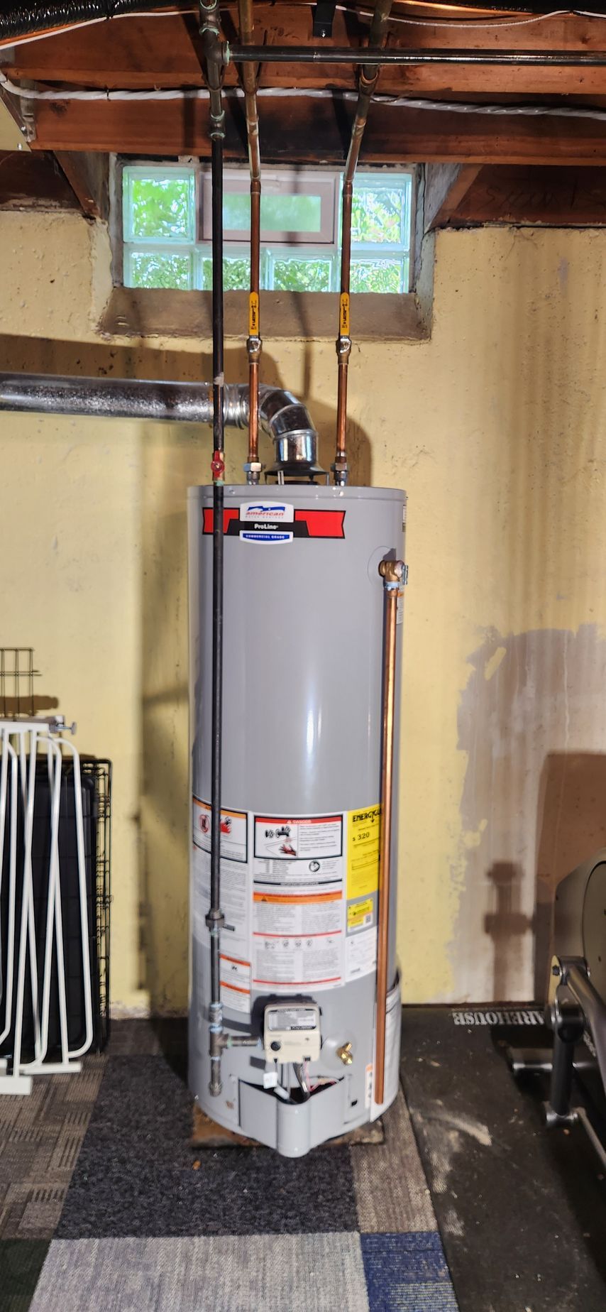 Water heater in a basement utility area with exposed pipes and a small window.