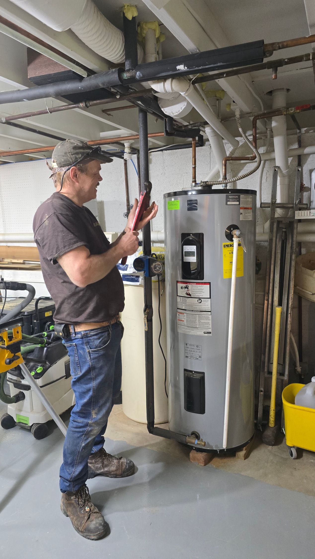 Technician inspecting a water heater and pipes in a utility room, using a tablet or phone.