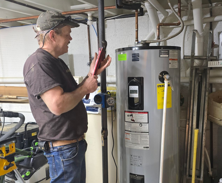 Technician inspecting a water heater in a basement utility room, holding a tablet.