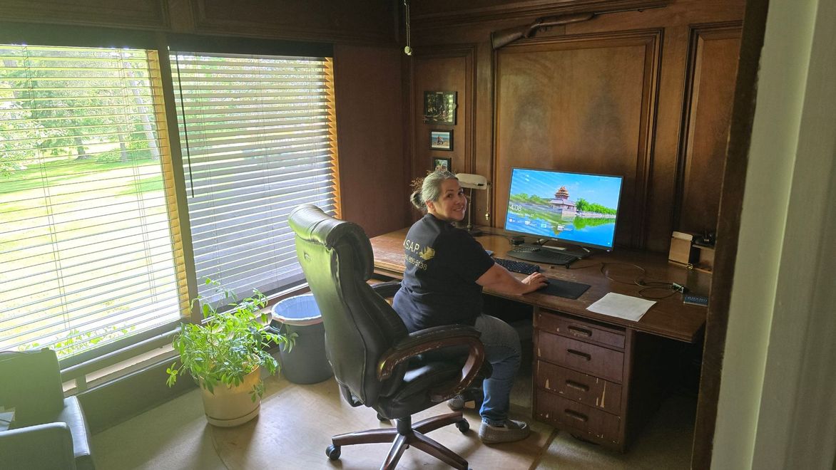 Person seated at a desk in a wood-paneled home office, working on a computer beside bright windows and plants