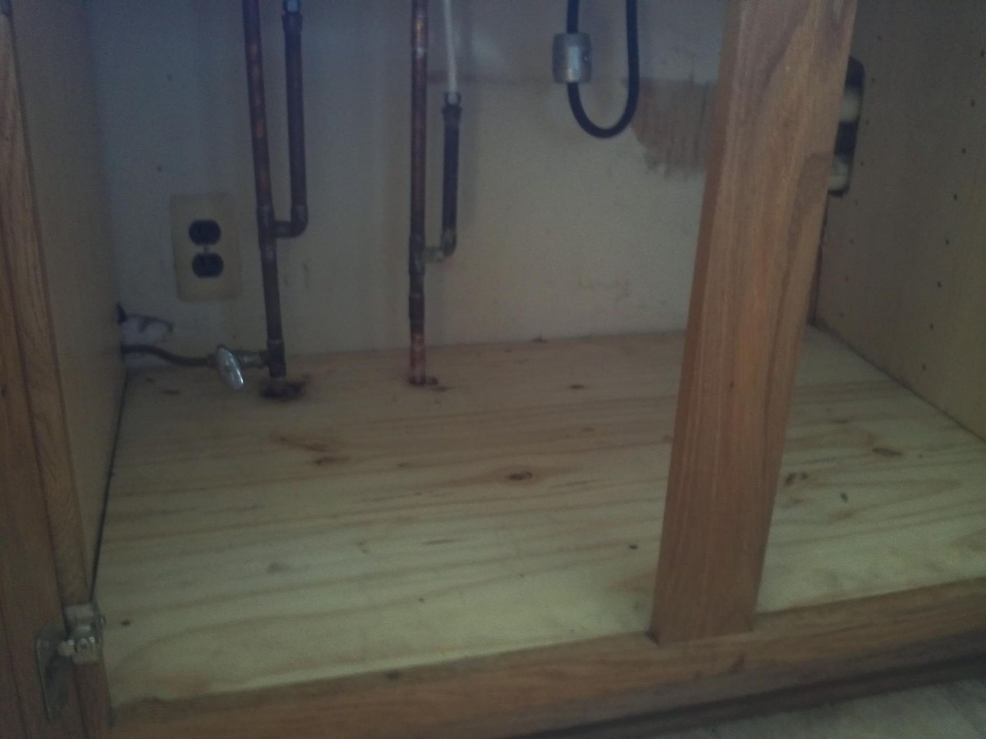Empty cabinet under a sink with exposed plumbing and a white electrical outlet on the left.
