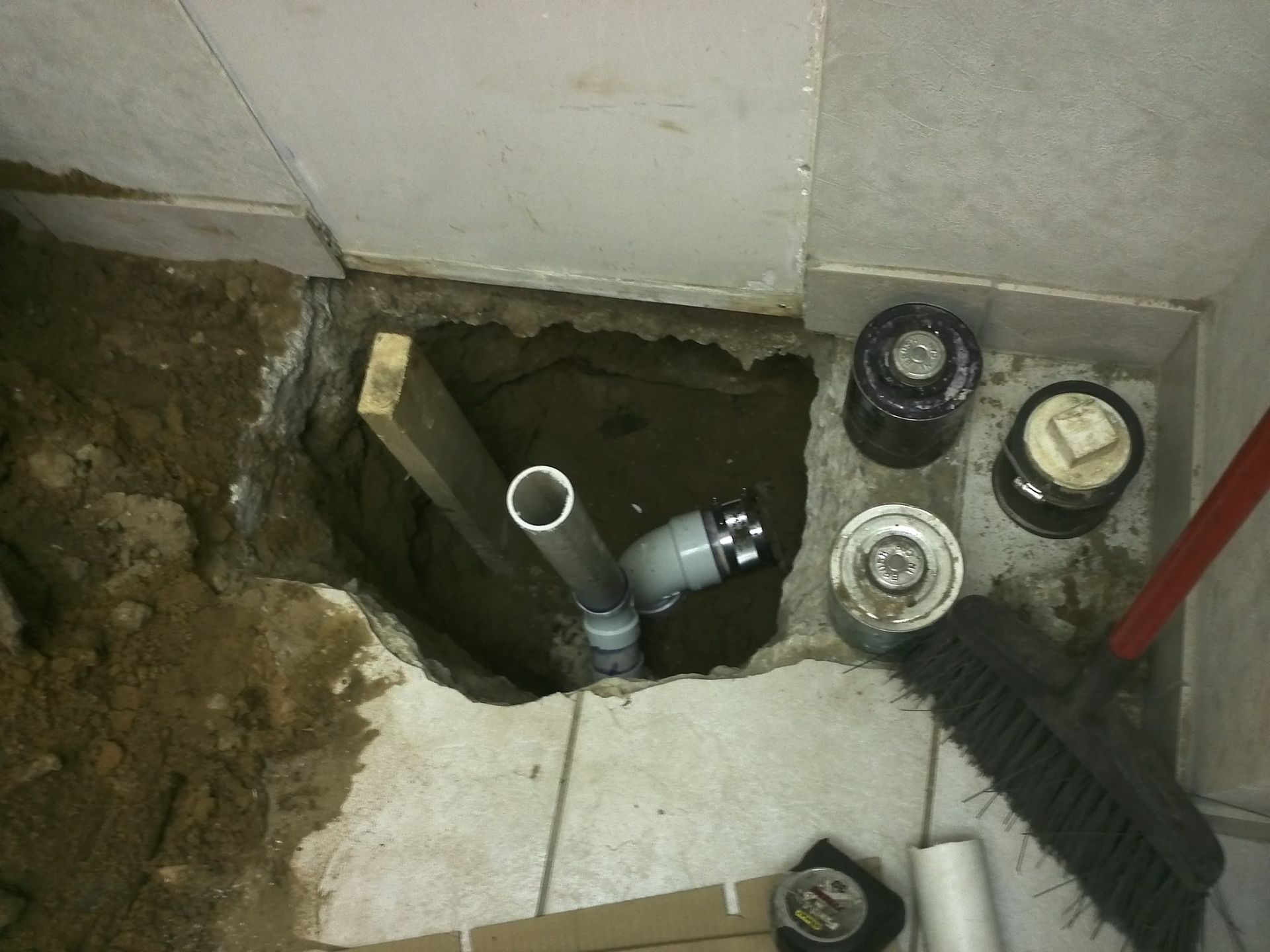 Open floor access hole exposing drain pipes beside bathroom tiles and inspection caps