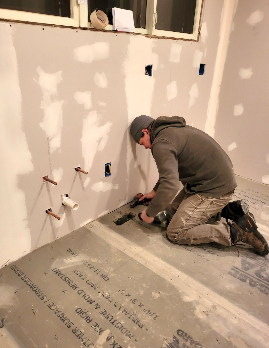 Worker kneeling to install baseboard in a room under renovation with exposed drywall patches