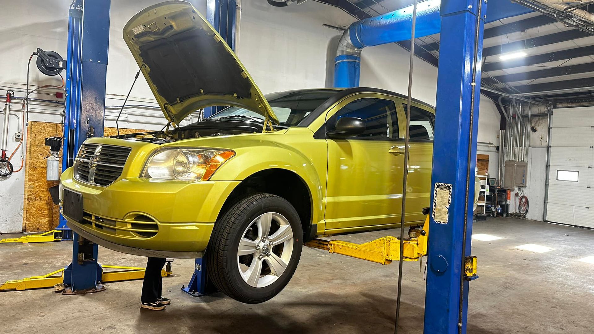 A yellow car with the hood up is on a lift in a garage.