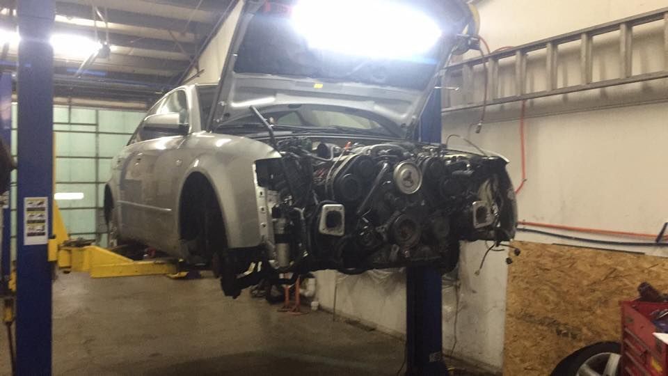 A car with the hood up is on a lift in a garage.