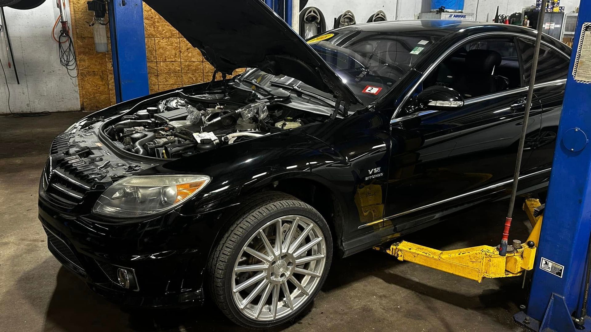 A black car with the hood up is on a lift in a garage.