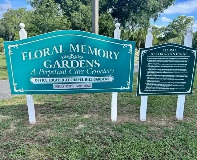 Sign for Floral Memory Gardens cemetery, teal with white trim, floral decoration guide to the right.